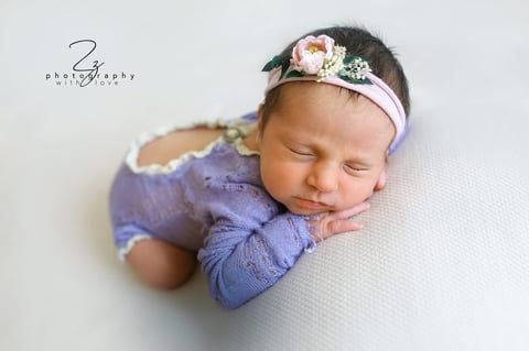 Newborn baby girl sleeping peacefully on white surface wearing purple knit outfit and floral headband