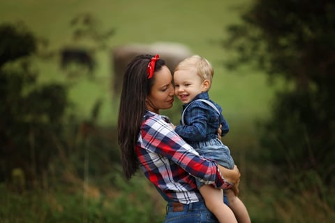 Woman in plaid shirt holding smiling toddler outdoors with green foliage background