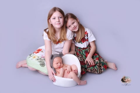 Two young girls sitting beside a newborn baby in a white bowl on a lavender background