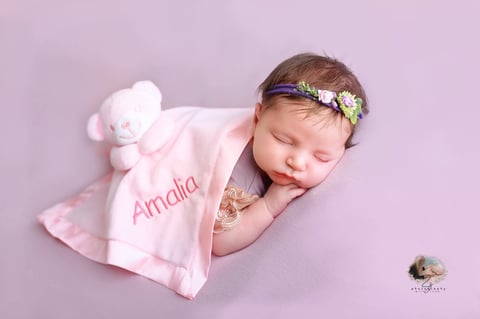 Sleeping newborn baby wearing a pink blanket and floral headband on a lavender background